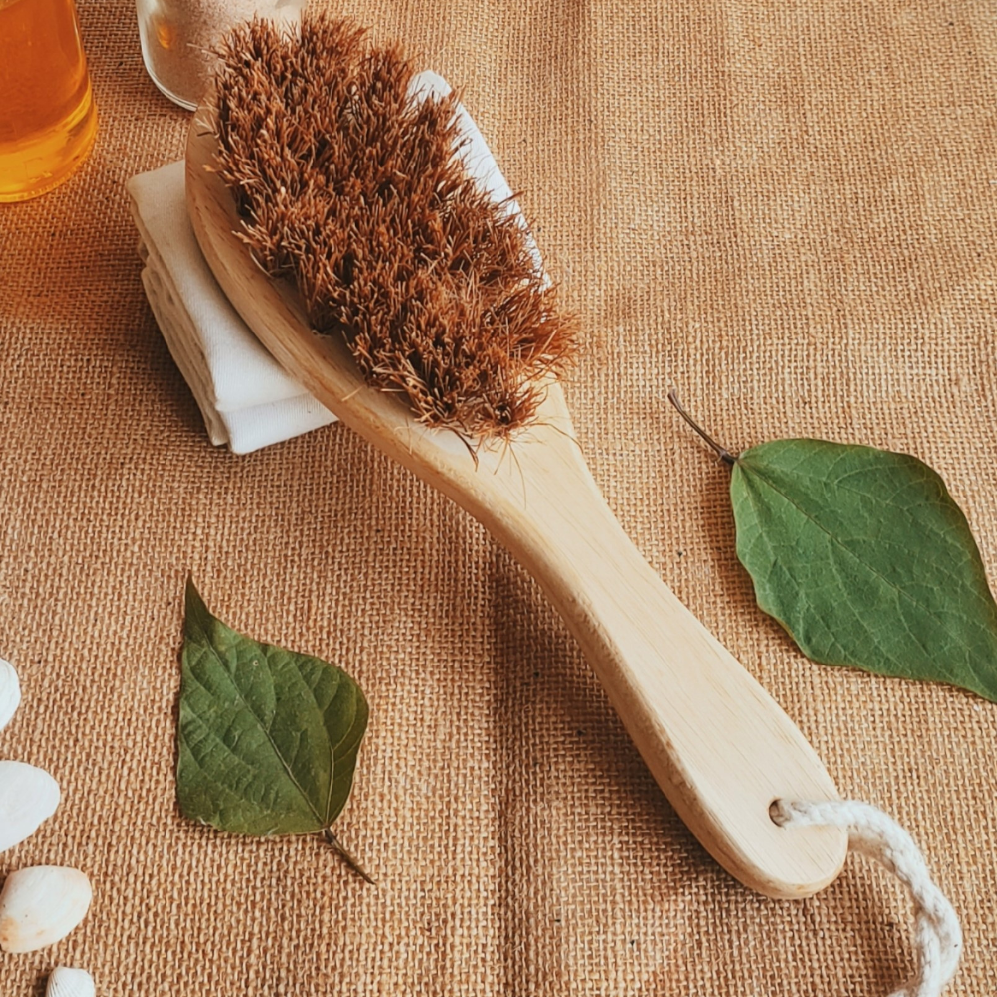 Top-down view of Almitra Sustainables’ coconut fiber dry body brush featuring a natural bamboo handle, placed on a jute fabric with green leaves, promoting zero-waste self-care and eco-conscious grooming.