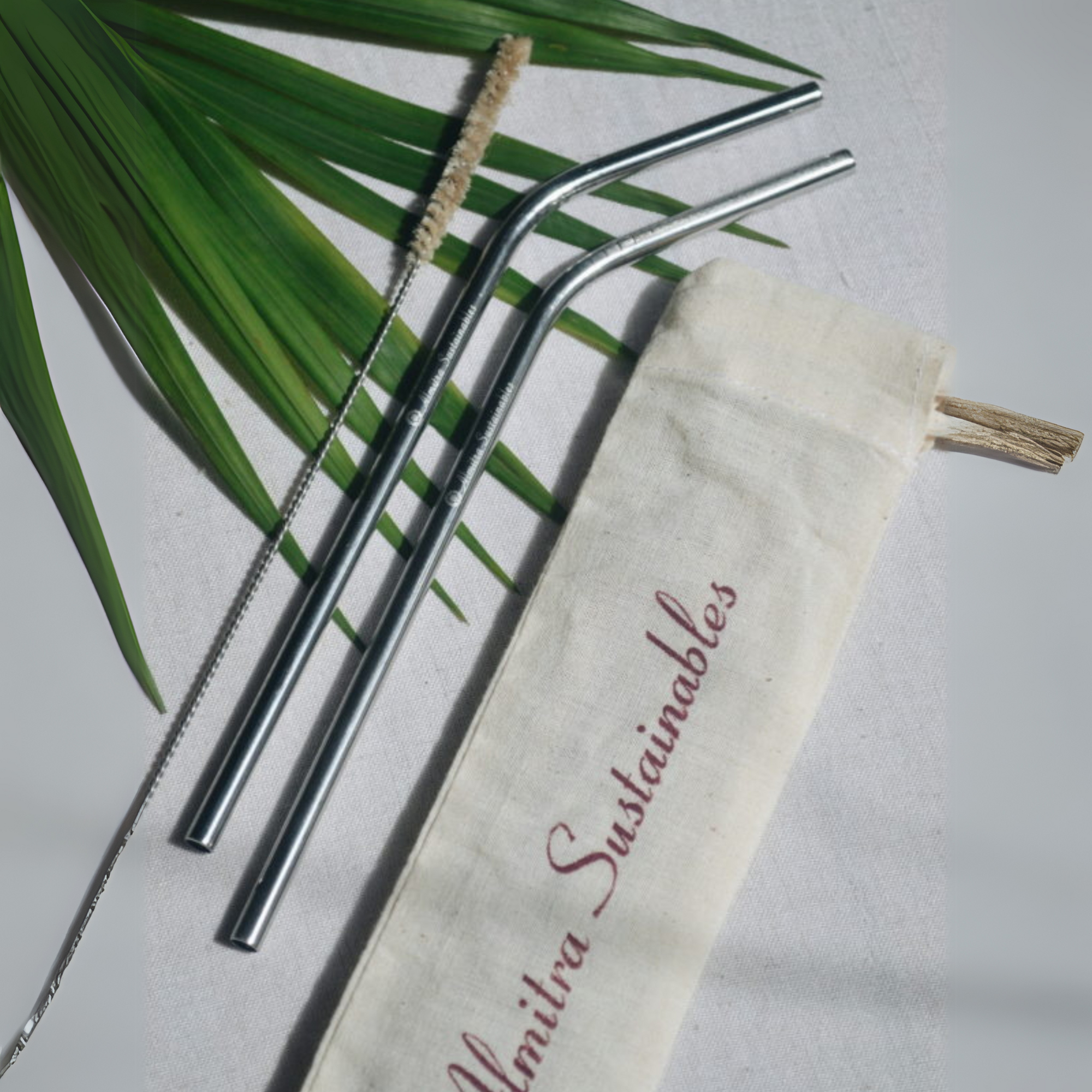 Set of two stainless steel bent straws with a natural bristle cleaning brush and cotton travel pouch printed with Almitra Sustainables branding, laid on a natural palm leaf backdrop – ideal for eco-conscious hydration on the go.